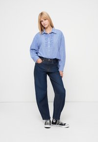 Μπλε ριγέ πουκάμισο σε υπερμεγέθη γραμμή με λοξές πιέτες στο μπροστινό μέρος, σε συνδυασμό με φαρδιά ψηλόμεσα σκούρα τζιν και μαύρα ψηλά αθλητικά παπούτσια. Απλό φόντο.