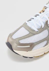 Sneaker léger avec une tige en maille blanche, des accents en cuir gris et une semelle en caoutchouc texturée. Présente des lacets blancs plats et des lignes de design.