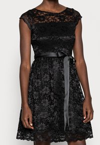 Robe en dentelle noire à manches courtes, corsage ajusté et jupe évasée. Dotée d'une ceinture en satin nouée à la taille et d'un ourlet festonné.