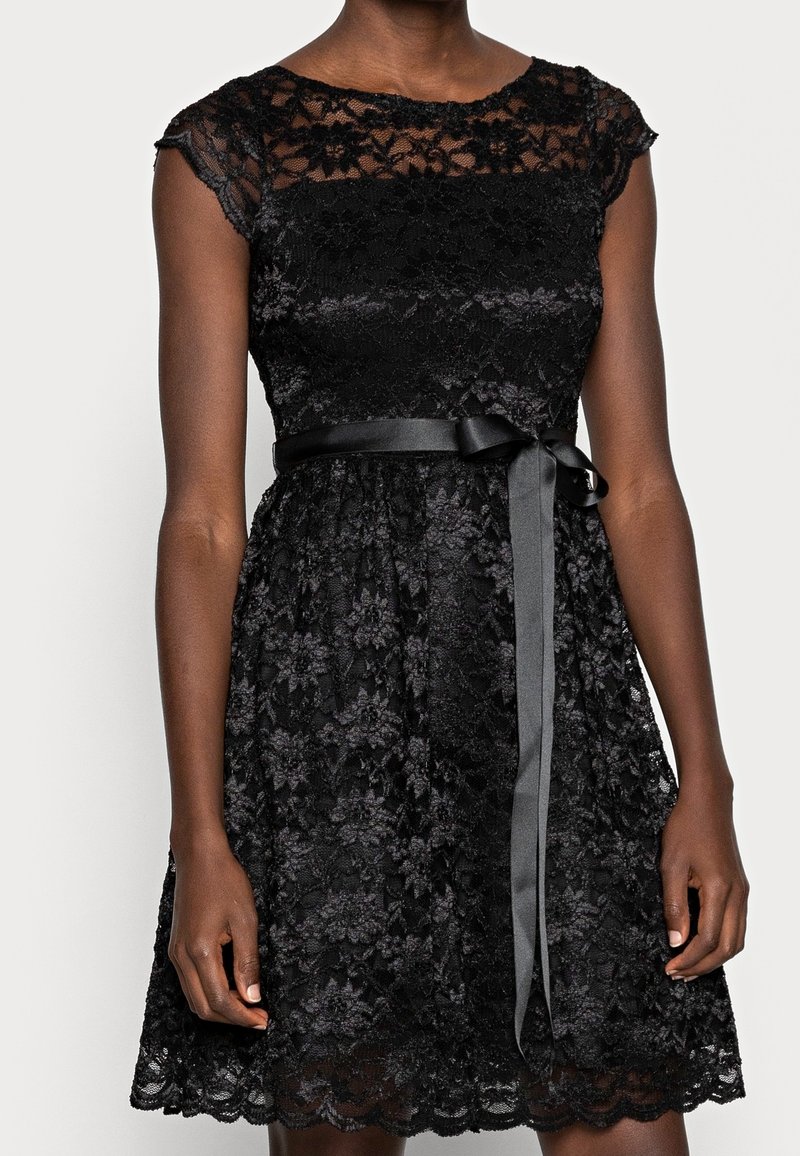 Robe en dentelle noire à manches courtes, corsage ajusté et jupe évasée. Dotée d'une ceinture en satin nouée à la taille et d'un ourlet festonné.