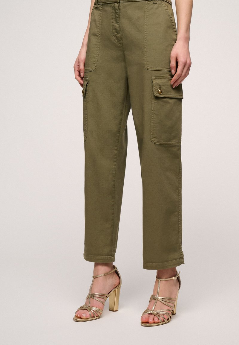 Pantaloni cargo verde oliva con vita alta, due tasche frontali e dettagli in bottone dorato, abbinati a sandali con tacchi alti e strappi dorati.