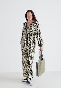 Robe longue à motifs noir et blanc avec des designs géométriques, associée à un sac fourre-tout vert et des baskets noires. Elle présente de longues manches et un décolleté en V.