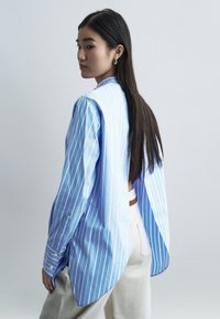 Νέα γυναίκα με μακριά σκούρα μαλλιά φοράει μπλε και άσπρο ριγέ φαρδιά πουκάμισα ανοιχτό στο πίσω μέρος, συνδυασμένο με εκρού παντελόνι σε απλό φόντο.