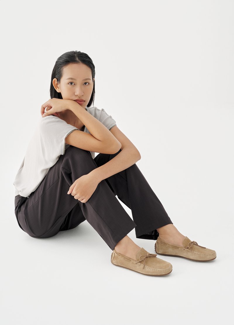 Jonge vrouw met zwart haar, witte t-shirt, zwarte broek en beige suède loafers, zittend op de grond met haar armen op haar knieën.