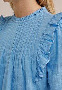 Svijetloplava bluzica s nabranim izrezom i rukavima, s teksturiranim vertikalnim plisiranim detaljima i mekanim, laganim materijalom.