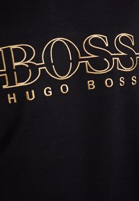 Tessuto nero con testo ricamato in oro che legge "BOSS HUGO BOSS" in lettere maiuscole grandi e più piccole.