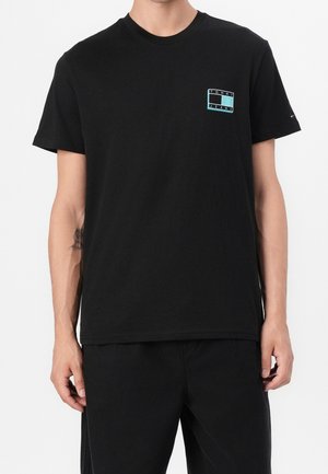 Mężczyzna noszący prostą czarną koszulkę z krótkim rękawem z małym prostokątnym logo Tommy Jeans na lewej piersi.