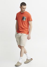 Camiseta roja de manga corta con un diseño de bolsillo con hojas, combinada con pantalones cortos beige. Modelo lleva calcetines blancos y zapatos de hombre de color marrón claro.