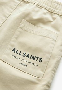 Beige bomuldsbukser med elastisk talje, med en syet lomme med teksten "ALLSAINTS" i sort, og en glat tekstur.