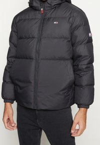 Sort puffjakke med hætte, featuring en frontlynlås, elastiske manchetter og en logo-patch på venstre skulder. Glat, skinnende materiale.