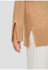 Maglione beige con orlo a coste e spacco laterale; realizzato in un morbido tessuto testurizzato. Mano mostrata appoggiata sul capo.