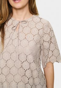 Top in pizzo beige con motivi floreali, maniche corte e scollatura con fiocco. Il tessuto presenta una texture delicata e semi-trasparente.