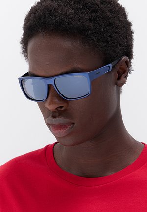 Primo piano di una persona che indossa occhiali da sole rettangolari blu e una camicia rossa, che guarda verso il basso su uno sfondo semplice.