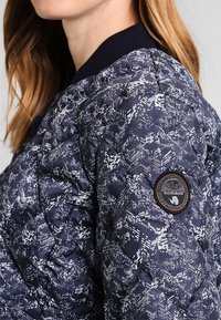 Gestepptes Marine-Jackett mit weißen geometrischen Mustern, geripptem Kragen und einem runden Stoffpatch am linken Arm. Strukturierte Oberflächenstruktur sichtbar.