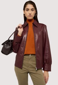 Burgunderfärgad läderjacka med dragkedja, spetsig krage och ballongsärmar. Bärs över en orange polotröja och matchas med olivfärgade byxor.