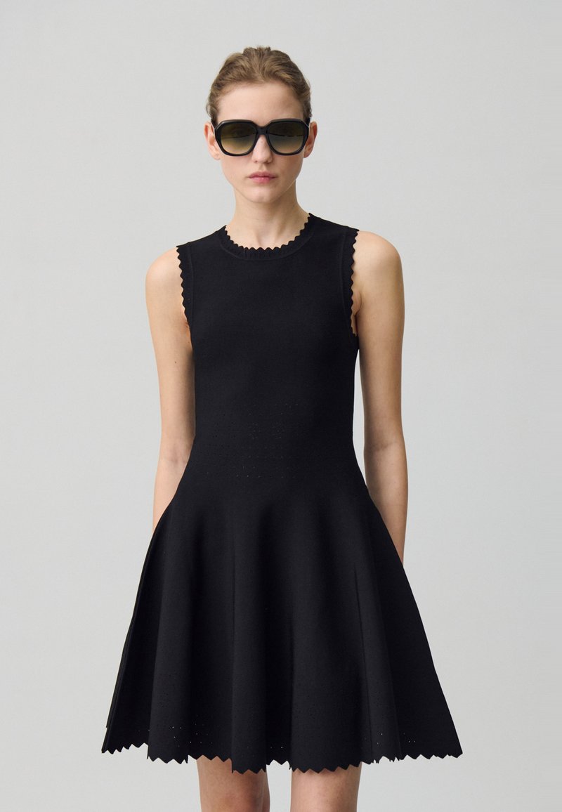 Robe noire sans manches en tissu texturé, avec des bords festonnés, un corsage ajusté et une jupe évasée. Des perforations notables à travers toute la pièce.