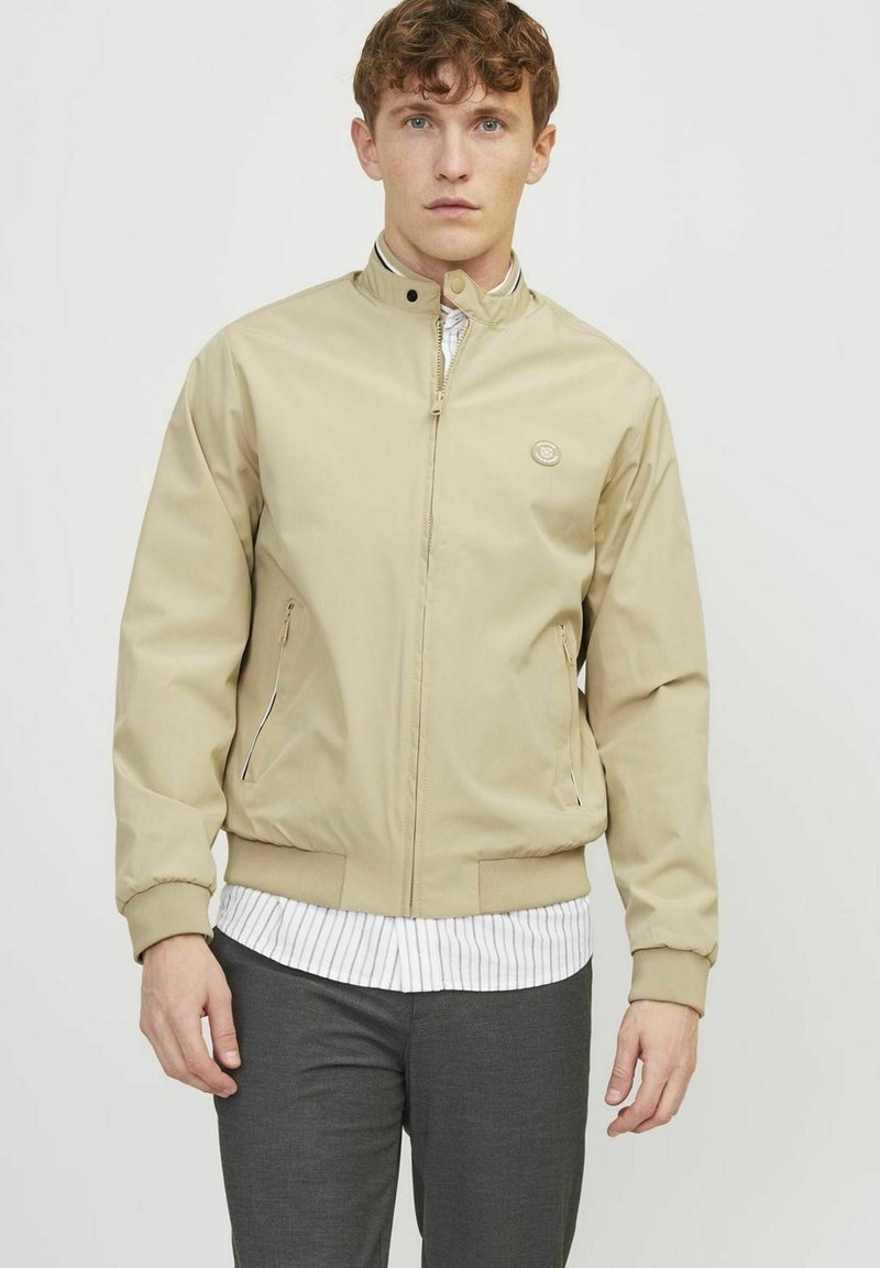 Veste beige à fermeture éclair avec col montant, dotée de poches latérales, de poignets et d'un ourlet côtelés, et d'un petit logo sur la poitrine. Chemise rayée en dessous.