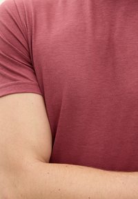 T-shirt rossa in cotone con collo girocollo e maniche corte, caratterizzata da una texture liscia e dettagli di cucitura sottili. Braccio parzialmente visibile.