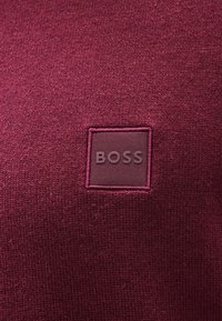 Μπλουζάκι σε βουργουνδί απόχρωση με τετράγωνο λογότυπο "BOSS" που είναι ανάγλυφο σε πιο ανοιχτή απόχρωση, περιτριγυρισμένο από μια υφασμάτινη μπορντούρα.