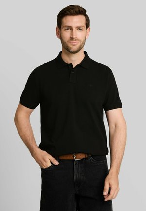 Man with short brown hair and light beard wearing a black short-sleeve polo shirt, black jeans, and a brown belt against a gray background.