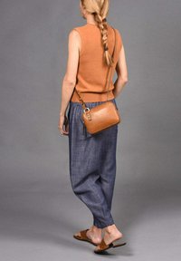 Suéter sin mangas marrón, pantalones sueltos azules, bolso cruzado de cuero beige y sandalias planas marrones. Un fondo neutro resalta el conjunto.