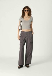 Mujer con cabello rizado, usando gafas de sol, un top corto gris, pantalones cargo de pierna ancha, sandalias de plataforma negras y llevando un bolso plateado al hombro.