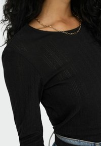 Schwarzes Langarmtop mit strukturiertem Rippdesign. Das Shirt hat einen runden Ausschnitt und wird mit Jeans kombiniert, wobei Hautpartien und eine goldene Kette betont werden.
