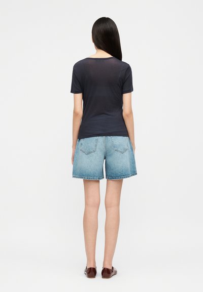 Vrouw staand, met de rug naar de kijker toe, gekleed in een effen donkerblauw T-shirt met korte mouwen, lichtblauwe spijkershorts en donkerbruine platte schoenen.