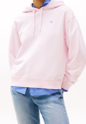 Persoon draagt een lichtroze hoodie over een blauw-wit gestreept shirt en lichtblauwe spijkerbroek, staand met handen in de zakken.