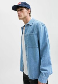 Lichtblauw denim overhemd met een klassieke kraag, knoopsluiting aan de voorkant, één borstzak en lange mouwen. Draagt over een wit tanktop.