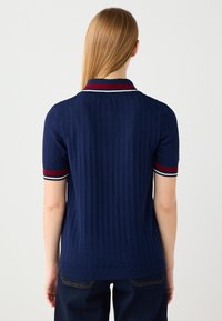 Polo shirt blu lavorato a maglia con maniche corte, caratterizzato da un colletto a coste accentuato da strisce rosse e bianche. Design testurizzato sul tessuto.