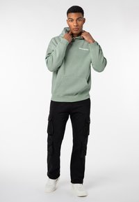 Helle grüne Kapuzenjacke mit großer Fronttasche, gerippten Bündchen und dem Text "WORLDWIDE". Schwarze Cargo-Hosen mit mehreren Seitentaschen und weißen Sneakers.