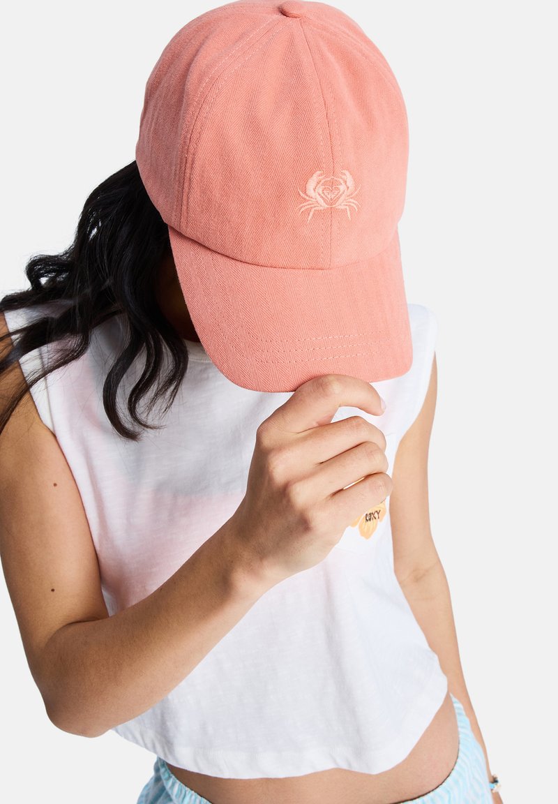 Gorra rosa de espiga con un bordado de cangrejo en el frente, sostenida por una persona que lleva un top blanco corto. La gorra tiene una visera curva.