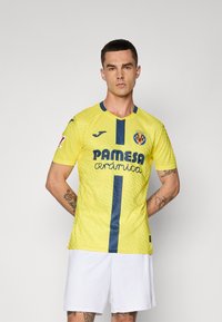 Mees, kes kannab kollast Villarreal CF jalgpallivormi sinise vertikaalse triibuga ja valgeid lühišortse, seismas halli ühetoonilise tausta ees.
