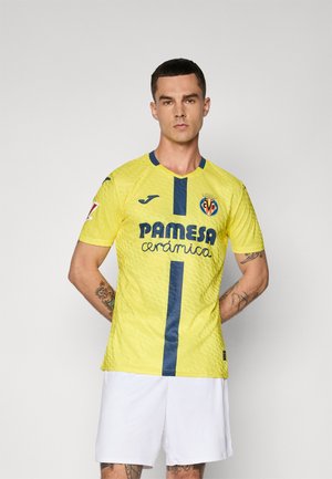Muž v žltej futbalovej dresu Villarreal CF s modrou vertikálnou pruhom a bielych šortkách, stojaci na obyčajnom šedom pozadí.