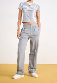 Persona vestida con un top corto a rayas blancas y azul marino, pantalones plegados gris claro, zapatillas blancas, con un tatuaje en el brazo izquierdo, de pie sobre un suelo amarillo.