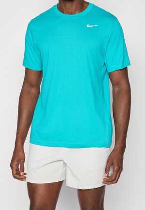 Homme portant un t-shirt Nike turquoise vif à manches courtes et un short blanc texturé sur un fond uni.