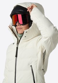 Biała ocieplana kurtka narciarska z wysokim kołnierzem, czarnymi zamkami błyskawicznymi i bocznymi kieszeniami. Posiada kaptur i jest noszona z okularami narciarskimi.