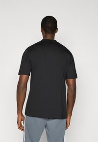 Μαύρο T-shirt με κοντά μανίκια και στρογγυλή λαιμόκοψη, κατασκευασμένο από λείο ύφασμα. Διαθέτει ίσια μέση και συνδυάζεται με γκρι παντελόνι φόρμας.