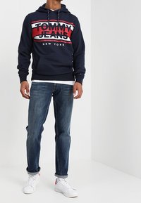 Sudadera azul marino con el logo "TOMMY JEANS" en rojo y blanco, combinada con jeans de lavado medio y zapatillas blancas. La sudadera tiene un bolsillo canguro.
