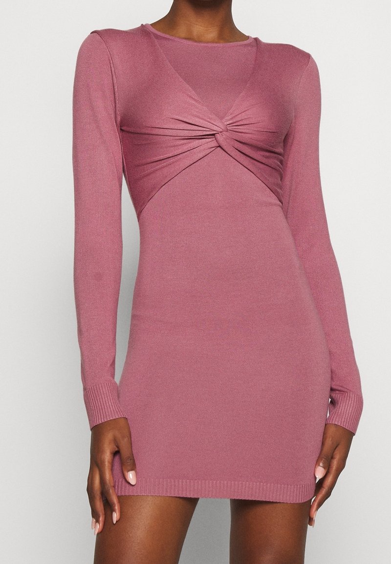Šaty s dlouhým rukávem, přiléhavé, v tlumené růžové barvě. Obsahují design s otočeným předním dílem a žebrovanou texturu na manžetách a lemu.