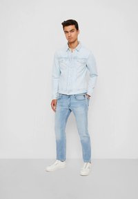 Ljusblå denimjacka med bröstfickor, knäppning och matchande ljusblå jeans. Modellen bär vita sneakers.