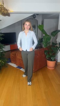 Πουκάμισο με κουμπιά σε ανοιχτό μπλε και λευκές ρίγες, παντελόνι ελιάς και γκρι αθλητικά παπούτσια· καφέ υφασμάτινος καναπές και μεγάλο φυλλώδες φυτό στο φόντο.