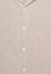 Chemise en lin beige avec un col revers et cinq boutons blancs à l'avant, présentant une texture subtile.