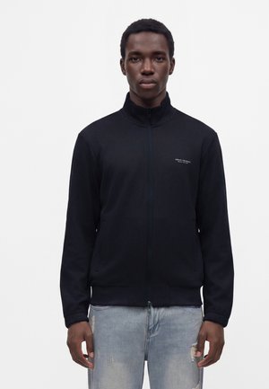 Junger Mann trägt eine schwarze Jacke mit Reißverschluss und kleinem Logo sowie hellblaue zerrissene Jeans und steht vor einem einfarbigen weißen Hintergrund.