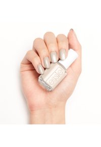 Bouteille de vernis à ongles Essie tenue dans une main. Le vernis est d'un beige clair scintillant, avec une bouteille en verre rectangulaire et un capuchon blanc. Ongles arrondis.