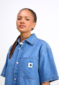 Denim shirt in light blue with short sleeves, button front, chest pocket, and black and white logo patch. Smooth texture with structured design.