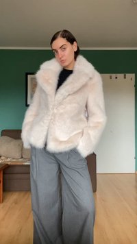 Veste en fausse fourrure rose avec un large col et une fermeture devant, associée à un pantalon gris loose, contre un fond intérieur simple.