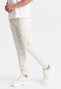 Pantalones de chándal color crema hechos de una tela suave, con un corte ceñido, piernas ajustadas y costuras en contraste, combinados con zapatillas blancas.