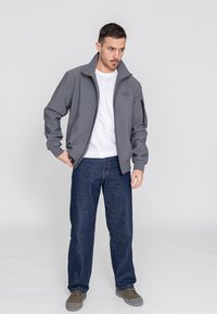 Graue Softshelljacke mit hohem Kragen und Reißverschlusstaschen, kombiniert mit blauen Jeans und grauem Schuhwerk. Einfaches, funktionales Design.